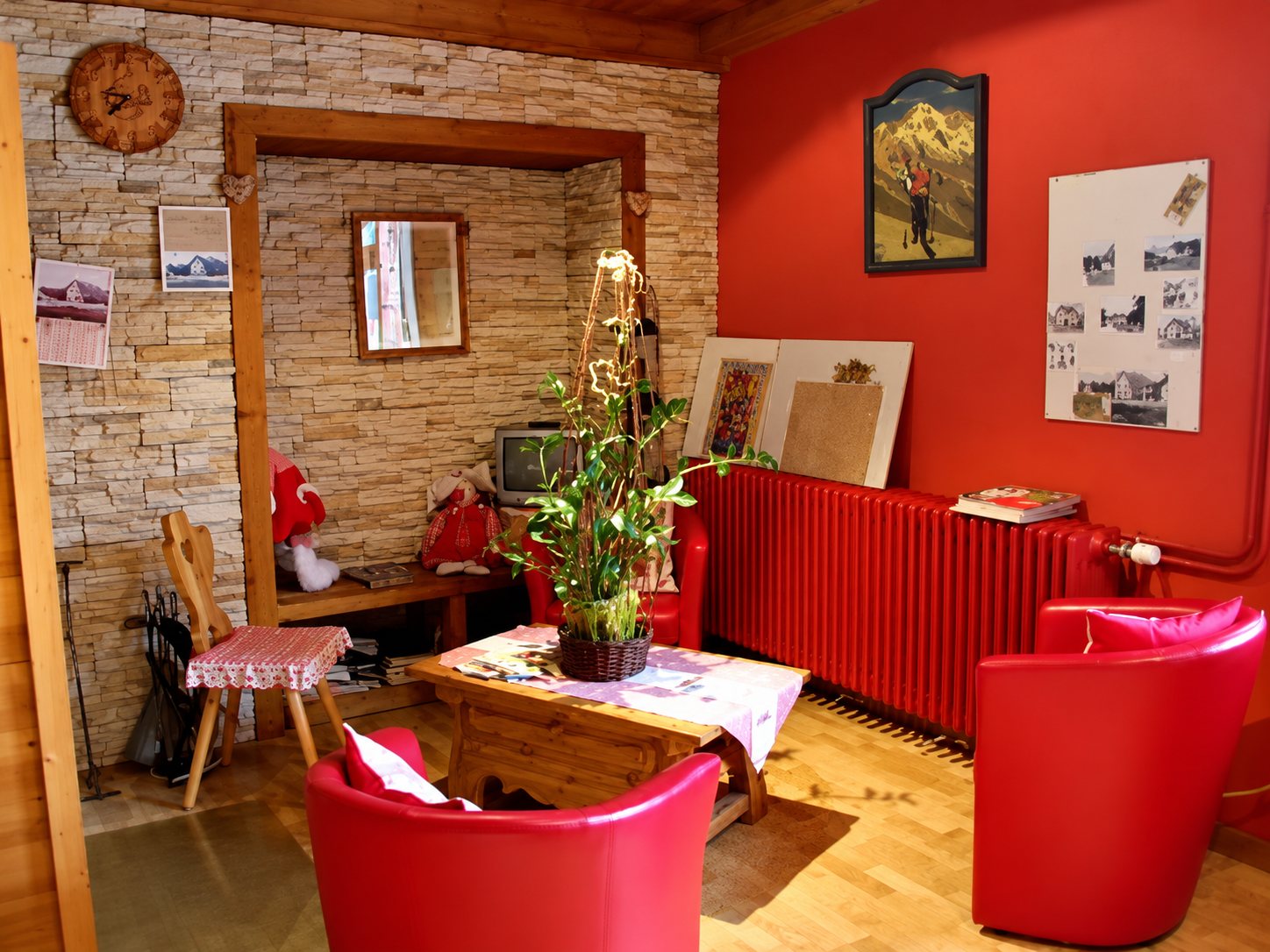 Un coin salon aux murs rouges et en pierre apparente, avec une table basse en bois, une plante verte, des fauteuils rouges et un radiateur rouge.