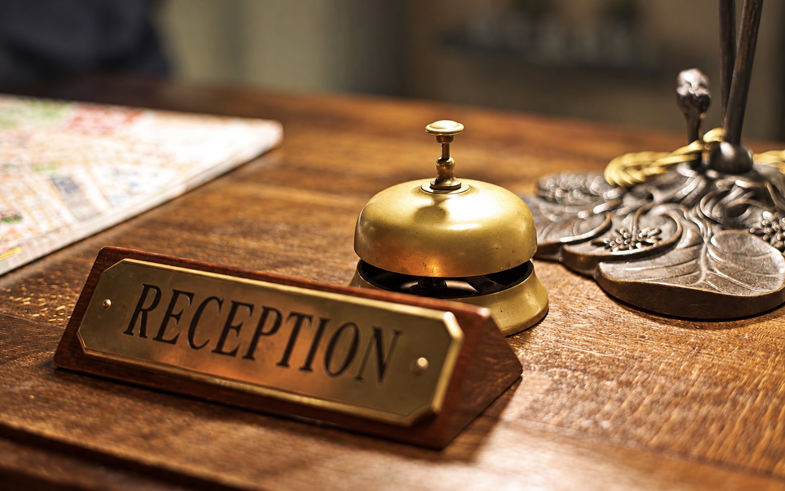 Une sonnette de réception en laiton et une plaque en laiton portant l’inscription Reception sont posées sur un comptoir en bois, à côté d’un pied de lampe ouvragé.