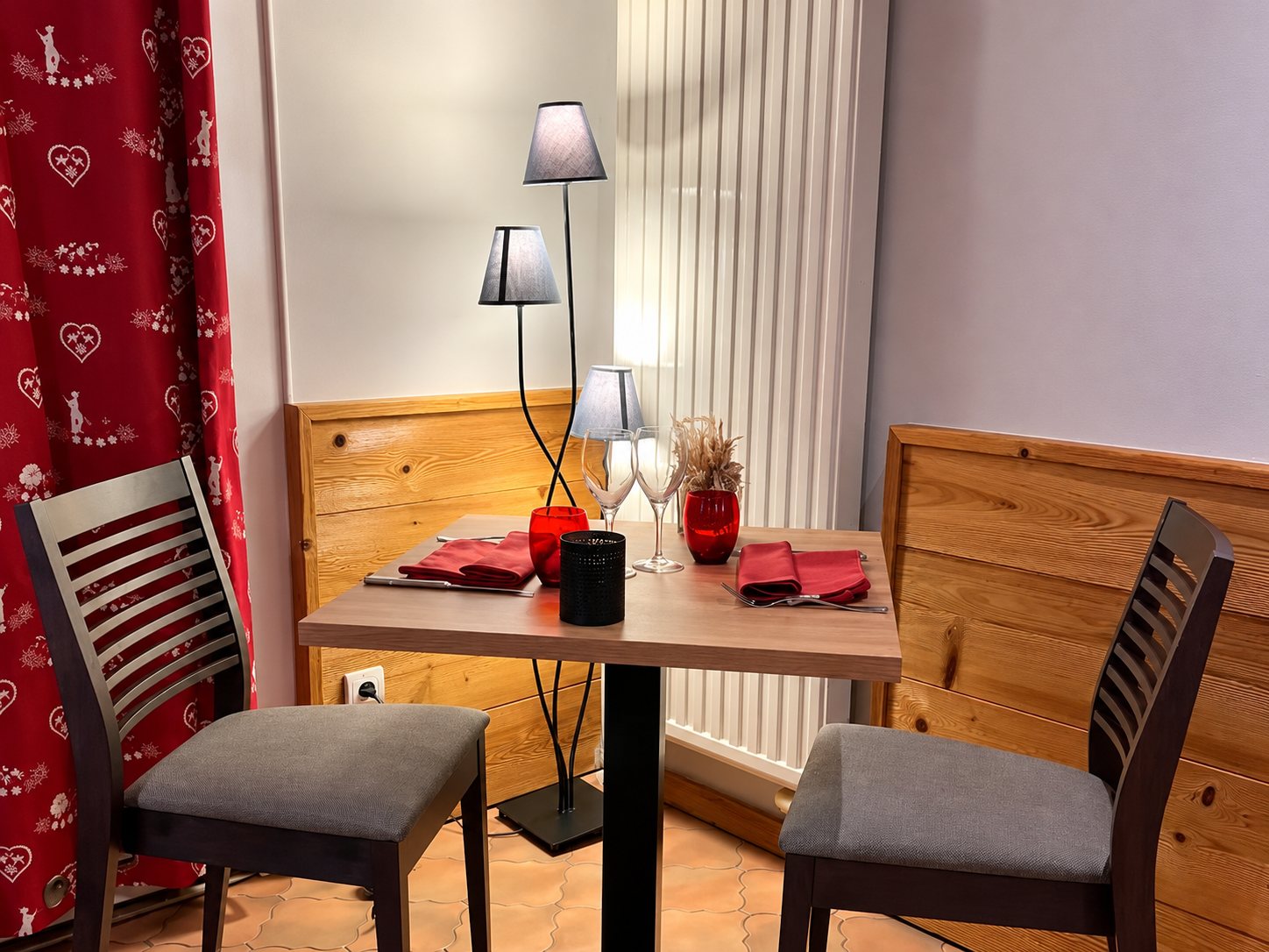 Une petite table dressée pour deux avec serviettes rouges et verres, entre deux chaises grises, devant des lambris en bois et un rideau rouge à motifs de cœurs.
