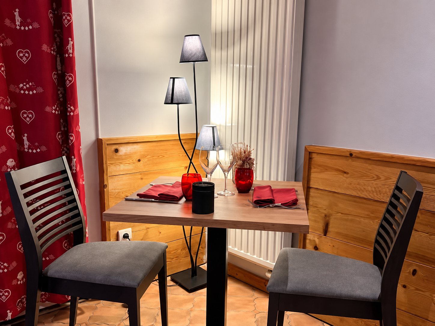 Une petite table en bois dressée pour deux avec serviettes rouges, verres et lampe, entourée de chaises grises et de boiseries.