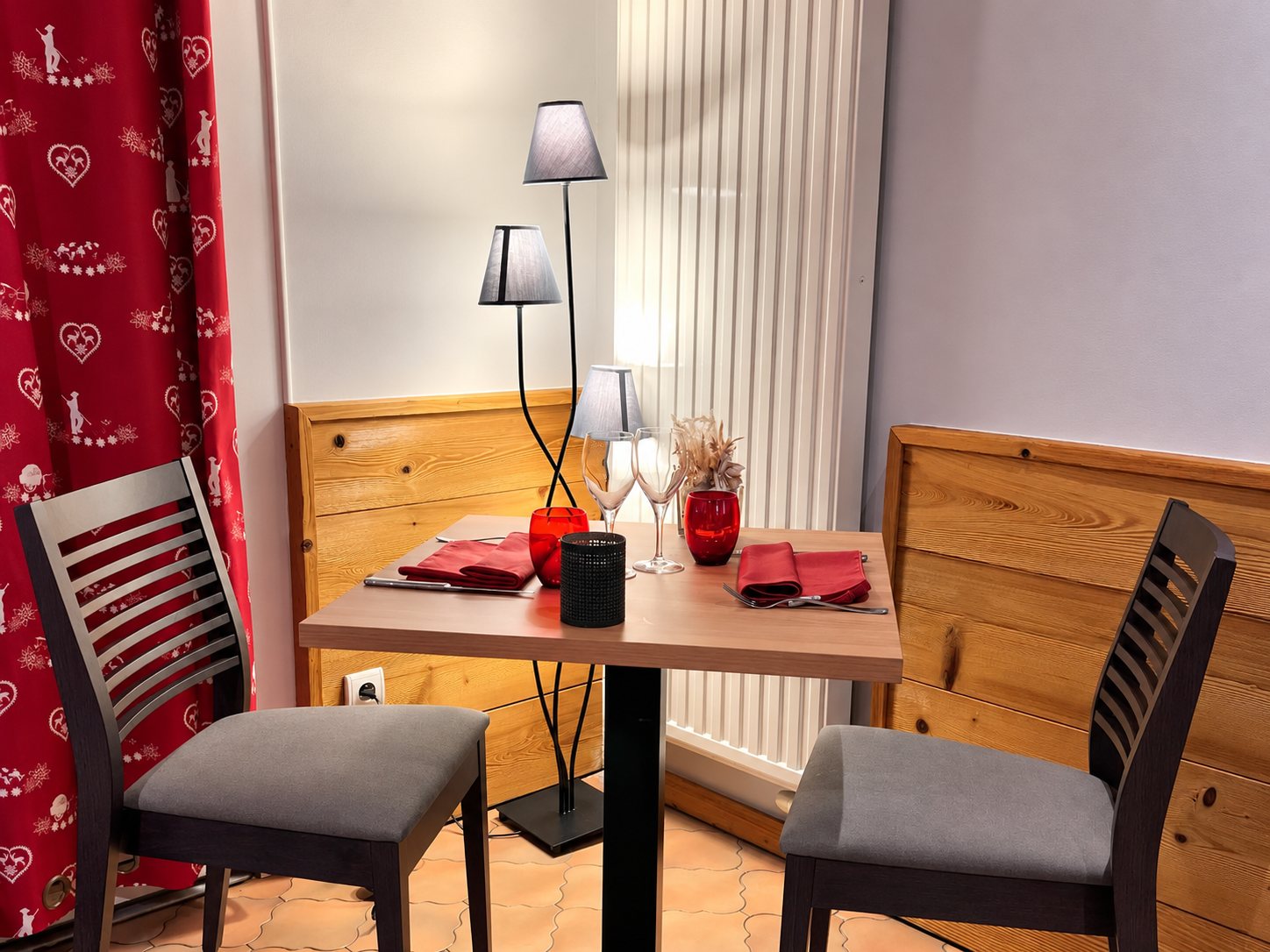 Une petite table carrée dressée pour deux avec serviettes rouges et verres, entourée de deux chaises, de lambris en bois, d’un lampadaire et d’un rideau rouge à motifs de cœurs.