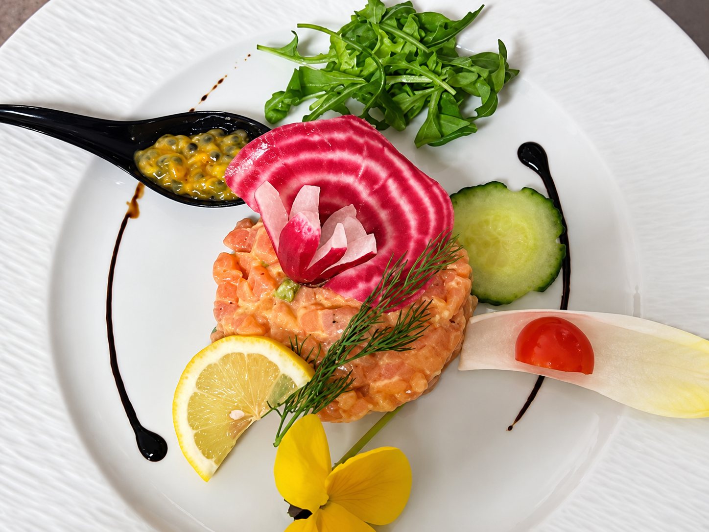 Assiette blanche garnie d’un tartare de saumon surmonté d’une betterave chioggia, accompagné de citron, concombre, roquette, fruit de la passion et fleurs jaunes.